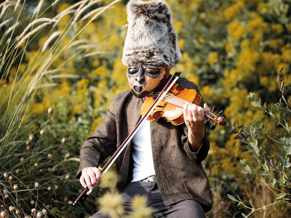 Eine Person mit einer Maske vor dem Gesicht und einem hohen Fellhut auf dem Kopf sitzt in einer Wiese und spielt auf einer Violine.