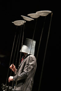 Bildbeschreibung: Mensch mit einem Eimer auf dem Kopf jongliert Teller auf langen Stöcken.