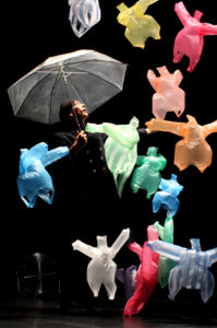 Bildbeschreibung: Ein Mann mit Regenschirm ist umringt von fliegenden bunten Plastiktüten.